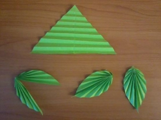 А теперь сделаем листочки: Берём лист зелёной бумаги, складываем по диагонали, обрезаем лишнее, складываем еще раз, затем разрезаем по сгибам. У нас получились треугольники. Складываем их гармошкой. Затем полученную форму складываем пополам, соединяя между собой длинную сторону.